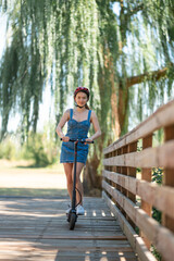 A young girl rides an electric scooter. Beautiful nature, a large wooden fence. Active rest, relaxation, tourism, environmentally friendly transport. Color image.