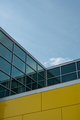 Exterior corner of a building with glass windows, a wall of yellow metal composite panels, a dark brown brick wall base with grey mortar. The background is a blue sky with white clouds on a sunny day.