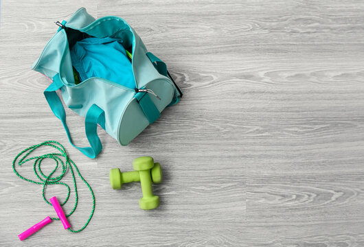 Bag with sportswear, skipping rope and dumbbells on light wooden background