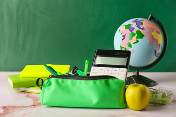 Pencil case with stationery, globe and apple on table in classroom