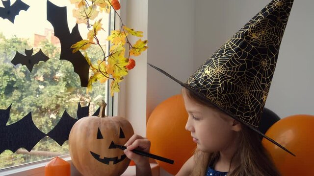 Preparation for Halloween: little girl paint funny face on pumpkin in witch hat with black marker. Room is decorated with bats, autumn leaves, balloons and candles. Holiday decoration concept