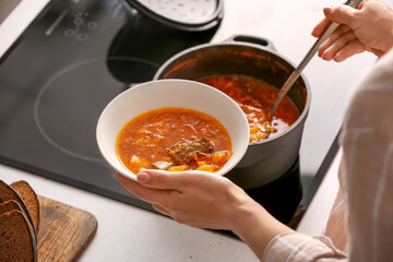 Woman pouring delicious borscht from cooking pot to bowl in kitchen