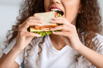 Tuinposter Young African-American woman eating tasty sandwich at home, closeup © Pixel-Shot