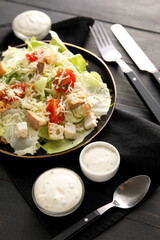 Plate of tasty Caesar salad bowls with sauce on black wooden background