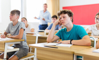 Teenager sitting in class room. Male teacher explaining something to them.