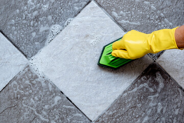 Top view of human hands wearing yellow rubber gloves are using a green color plastic floor scrubber to scrub the tile floor with a floor cleaner.