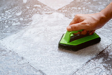 Close up human hand are using a green color plastic floor scrubber to scrub the tile floor with a floor cleaner.