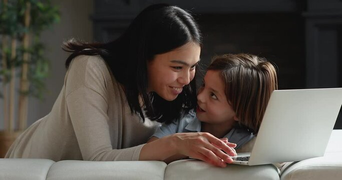 Caring young korean asian mommy or nanny teaching adorable small child boy using computer educational software applications, sitting together on comfortable sofa, online education or internet purchase