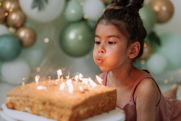 Little dark-skinned girl makes a wish and blows out the candles on the birthday cake. Cute girl...
