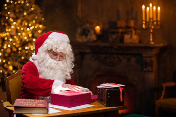 Workplace of Santa Claus. Cheerful Santa is conjuring over the gift box while sitting at the table. Fireplace and Christmas Tree in the background. Christmas concept.