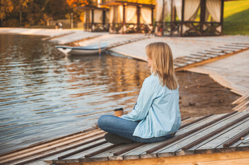 Autumn relax. A blonde woman sits on the shore of an autumn pond with a glass of hot coffee or tea in her hands. View from the back. A place for text.