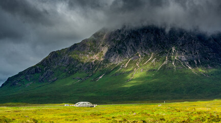 Along the West Highland Way. A bridge spans a river in the moor of the Glen Coe valley, at the foot of the Stob a Ghlais Choire. The sky is threatening and the sun plays on the walls of the mountain