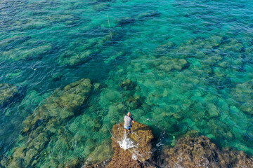 Fisherman throwing a fishing line while standing on a shallow lagoon rock, Aerial view.