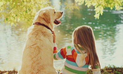 Little girl child with Labrador retriever dog sitting in sunny summer park near river