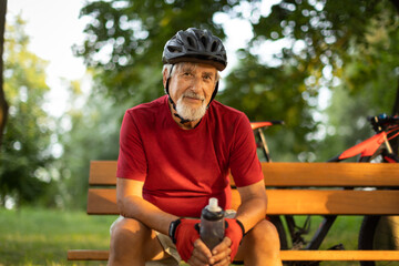 Senior man with his mountain bike going for a ride past the city limits in a lovely forest, getting the daily cardio dose, having a break on a bench