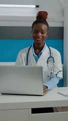 Portrait of healthcare specialist with white coat in cabinet at medical clinic. Young doctor using laptop, documents and tools on desk in healthcare office with modern equipment