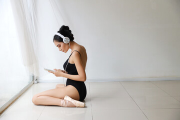 Fototapeta na wymiar Stunning shape of charming asian woman wearing black sportswear sitting with headphone. Beautiful girl listening music from mobile phone while training ballet dancing (technology and healthy concept)