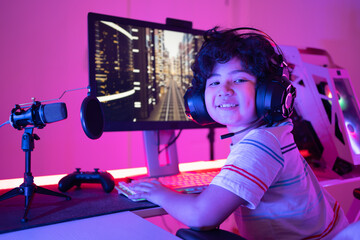 Cute curly-haired little Asian boy gamer in headphones sitting playing video games on PC with RGB...