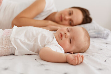 Indoor shot of attractive dark haired female wearing white t shirt lying on bed with her infant baby, having rest together, mommy looking at her cute daughter.