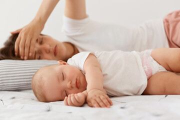 Tired exhausted female wearing white casual style t shirt lying near sleeping baby, keeping palm on...