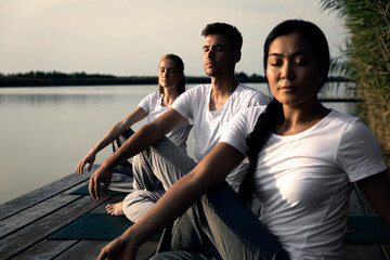 Group of people doing yoga exercises by the lake at sunset.