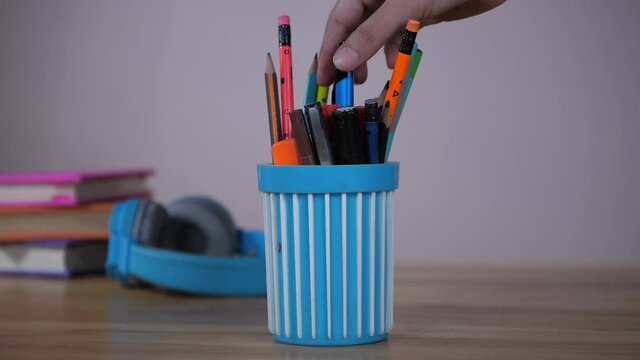 Pen stand with colored pens and pencils - A student taking a pen from a pen holder to write her notes. A few books and blue headphones kept together on a desk - study material  music  entertainment