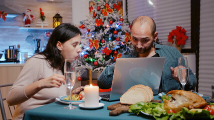 Festive couple looking at laptop and enjoying christmas dinner with technology. People eating chicken and drinking champagne while using device. Man and woman celebrating holiday.