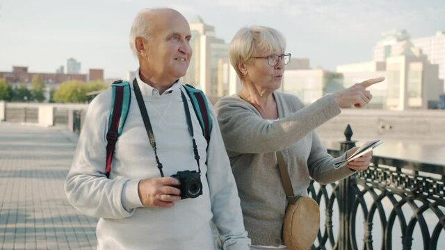 Senior tourists married couple walking in city holding map and photo camera talking laughing enjoying summer day outdoors. People and travelling concept.