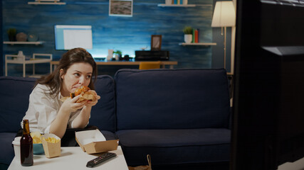 Caucasian woman eating hamburger from delivery bag while watching movie on television at home....