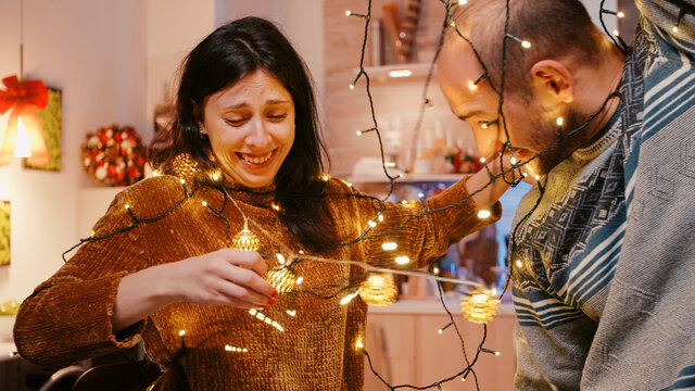 Couple Laughing And Getting Tangled In String Of Christmas Lights. Man And Woman Trying To Untangle Knot Of Garland With Twinkle Lights And Illuminated Bulbs. Holiday Decorations