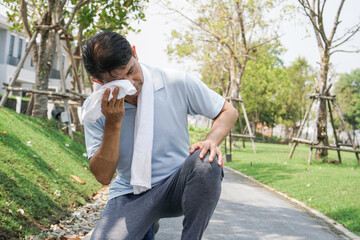 Asian senior tired man sitting and using towel to wipe a sweat while walking or jogging exercise at...