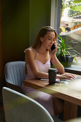 beautiful young asian woman sitting at coffee shop table surrounded by plants, working on laptop. beautiful lady calling by smartphone. modern online education, distant work, remote job, freelance