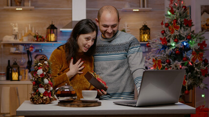 Cheerful couple receiving presents on video call for christmas celebration. Married people with gifts talking on online conference with family for holiday festivity in festive decorated kitchen