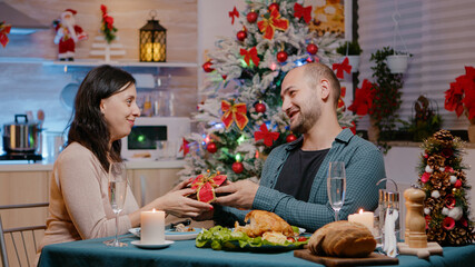 Couple celebrating christmas eve with presents while enjoying festive dinner. Man giving gift to cheerful woman and eating traditional food for holiday celebration. Seasonal festivity