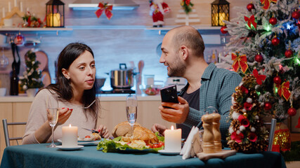 Couple celebrating christmas eve with festive dinner looking at smartphone for entertainment. Cheerful people enjoying traditional meal and alcohol for holiday celebration with technology