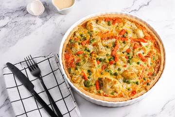 Baked french vegetable pie quiche filled with carrots, peas and cheese on a marble surface, cracked egg shell, flour, cream