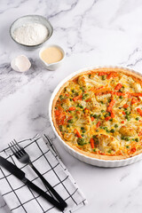 Baked french vegetable pie quiche filled with carrots, peas and cheese on a marble surface, cracked egg shell, flour, cream