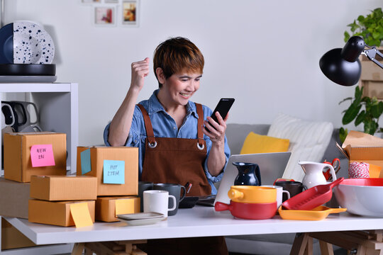 Successful Mature Asian Woman Entrepreneur, Business Owner With Arm Up Happy While Looking At Her Online Order From Smartphone. Online Selling Business Work At Home Concept