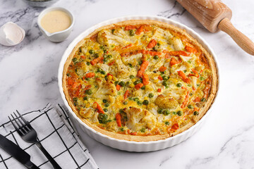 Baked french vegetable pie quiche filled with carrots, peas and cheese on a marble surface, cracked egg shell, flour, cream