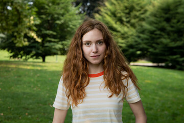 Cinematic shot of young happy red hair woman is smiling in camera in green city park. Concept of lifestyle, youth, diversity, women power, equality, femininity, women's day.