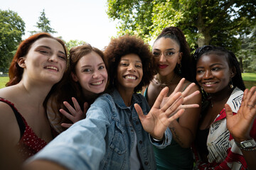 Cinematic shot of young happy girl friends of different ethnicities having fun to make selfie or technology video call in green city park. Concept of connection,friendship, lifestyle, youth,diversity.