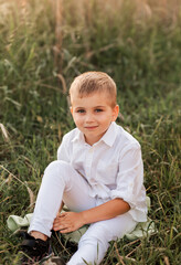 Naklejka na ściany i meble A small handsome boy blonde in a white shirt and white pants in nature in the summer. Happy childhood. Positive emotions