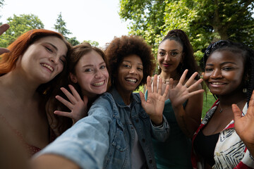 Cinematic shot of young happy girl friends of different ethnicities having fun to make selfie or technology video call in green city park. Concept of connection,friendship, lifestyle, youth,diversity.