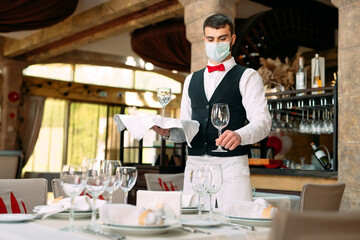A waiter in a medical protective mask serves the table in the restaurant.