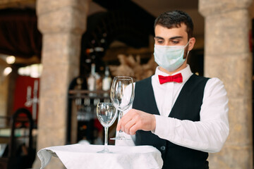 A waiter in a medical protective mask serves the table in the restaurant.