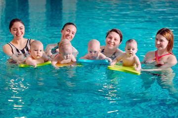 A group of mothers with their young children in a children's swimming class with a coach.