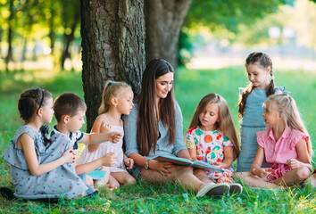 Children and education, young woman at work as educator reading book to boys and girls in park.