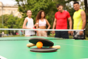 Friends talking near ping pong table outdoors, focus on rackets and ball