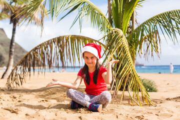 Little girl in santa hat sitting next to palm tree on beach