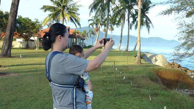 Local lifestyle Asian Chinese mother carry her baby while talking selfie photo with baby on beautiful tropical beach.
